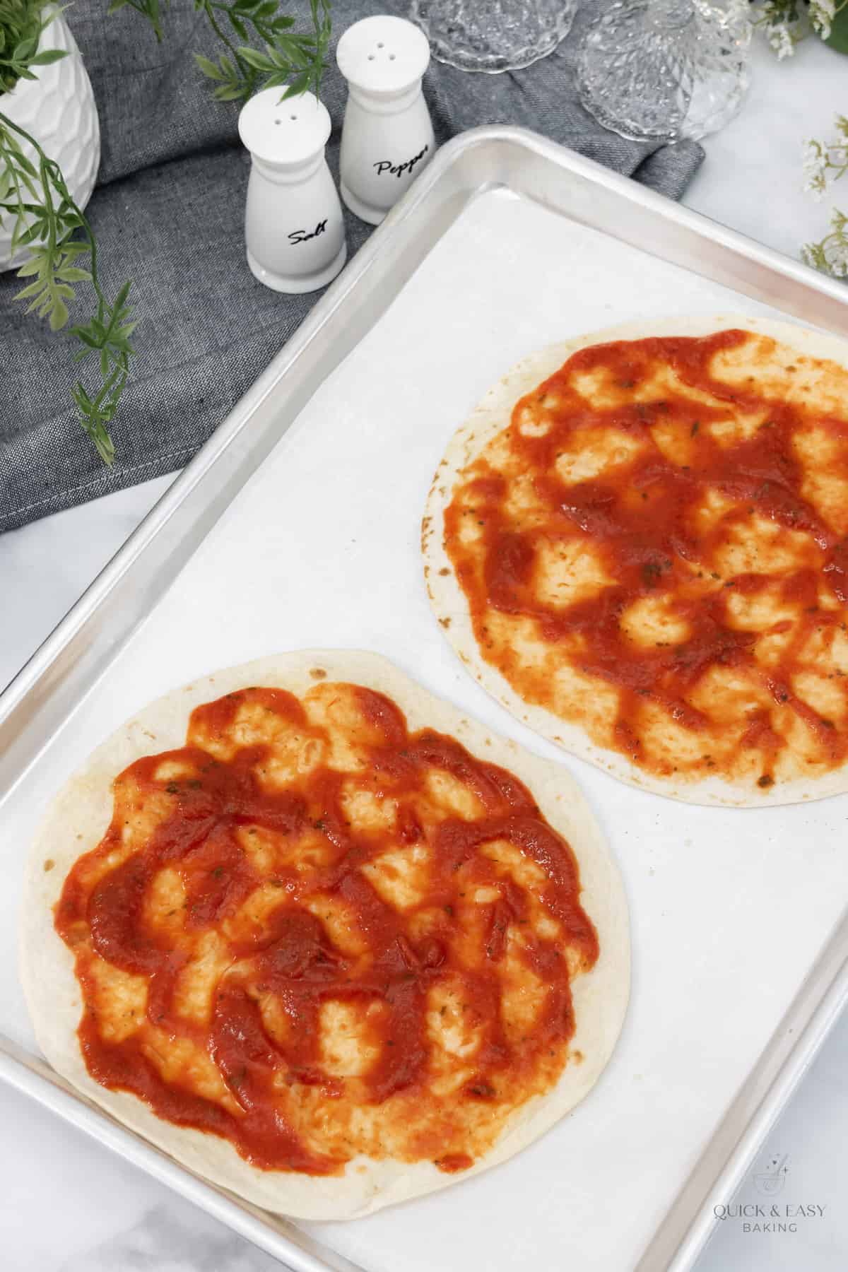 Pizza sauce spread onto tortillas on a baking sheet.